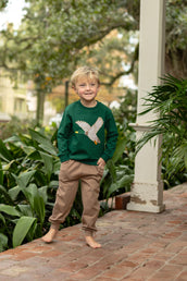 Young boy wearing a green sweater with a mallard design and brown pants standing on a brick path with plants in the background.